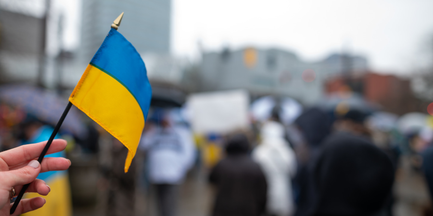 eine Hand mit einer Ukrainischen Flagge im Hintergrund stehen Menschen